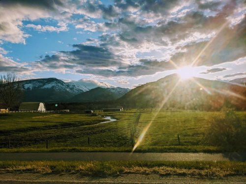 A scenic landscape featuring mountains, a field, and a bright sunset with clouds and a winding stream.