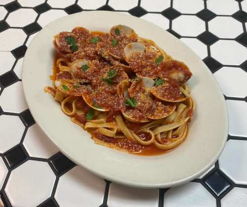 A plate of fettuccine pasta topped with clams, marinara sauce, and grated cheese, served on a black and white tiled surface.