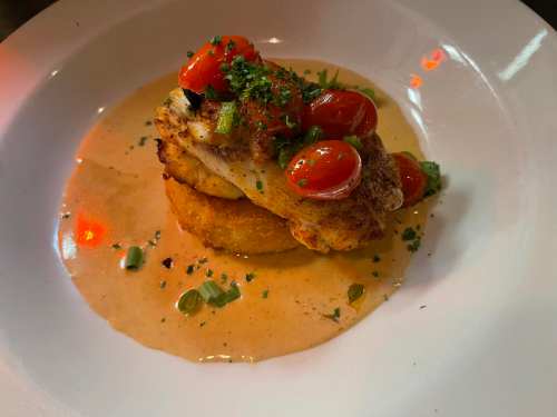 A plated dish featuring fish atop a fried cake, garnished with cherry tomatoes and herbs in a creamy sauce.