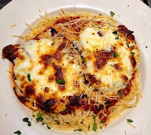 A plate of spaghetti topped with two pieces of cheesy chicken, garnished with parsley and grated cheese.