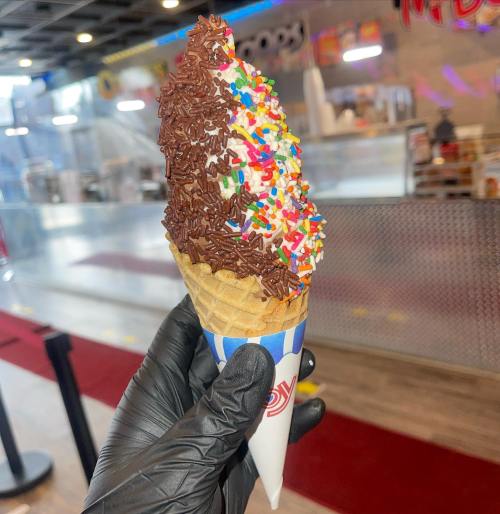 A hand in a black glove holds an ice cream cone topped with chocolate sprinkles and colorful confetti sprinkles.