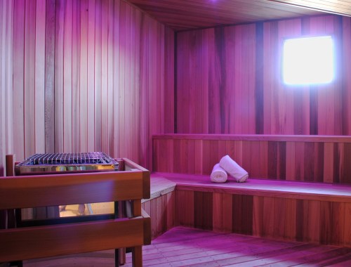 A wooden sauna interior with purple lighting, featuring benches and a small window. Two rolled towels are on the bench.
