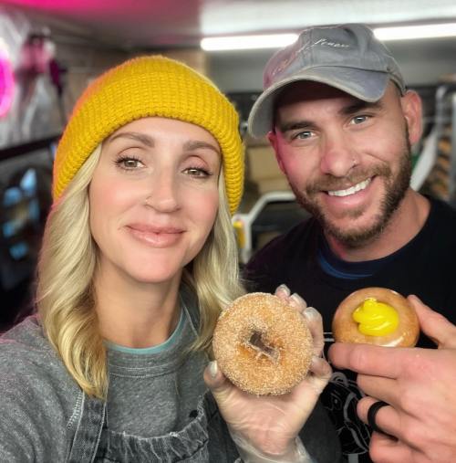 A smiling couple holds donuts, one with a yellow filling, in a cozy, brightly lit setting.