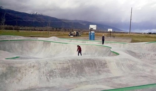 Two people skateboarding in a concrete skatepark surrounded by mountains and a basketball court in the background.