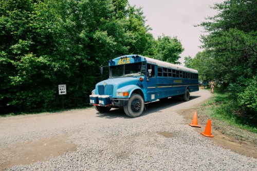 A blue school bus drives down a gravel road surrounded by green trees, with orange cones marking a one-way sign.