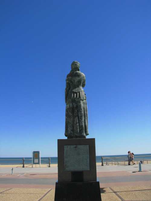 A statue of a woman stands facing the ocean under a clear blue sky, with people walking along the promenade in the background.