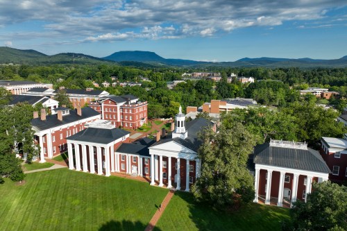 Aerial view of a college campus with red brick buildings, green lawns, and mountains in the background under a blue sky.