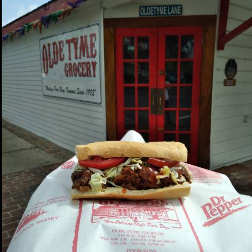 A delicious po' boy sandwich with lettuce and tomatoes, held in front of Olde Tyme Grocery's red door.