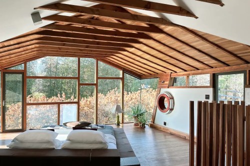 Cozy bedroom with wooden beams, large windows, and a view of greenery, featuring a bed and decorative plants.