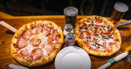 Two pizzas on a wooden table: one with pepperoni and cheese, the other with vegetables and olives, alongside drinks.