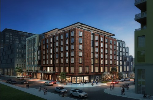 A modern brick building at dusk, featuring large windows and surrounding urban architecture, with cars and pedestrians nearby.