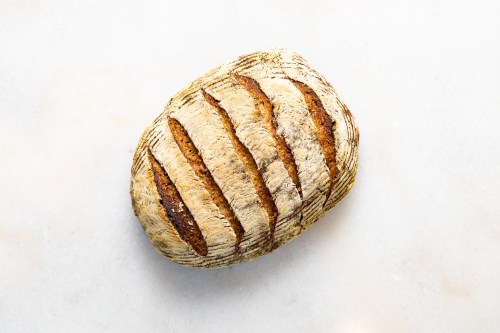 A round, crusty loaf of bread with scored lines on top, resting on a light marble surface.