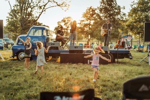 Children dance in a grassy field during a sunset concert, with musicians performing on stage and a vintage truck nearby.