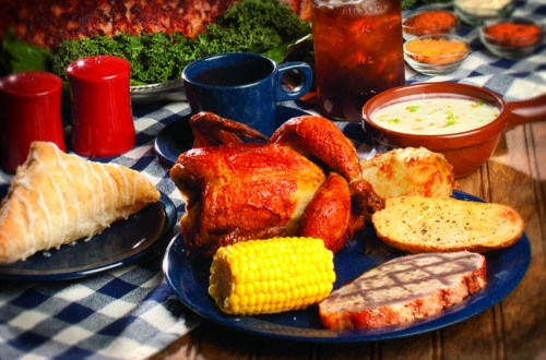 A hearty meal featuring roasted chicken, corn, bread, and soup, served on a blue plate with drinks in the background.