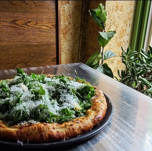 A freshly baked pizza topped with arugula and grated cheese, set on a table near a window with plants.