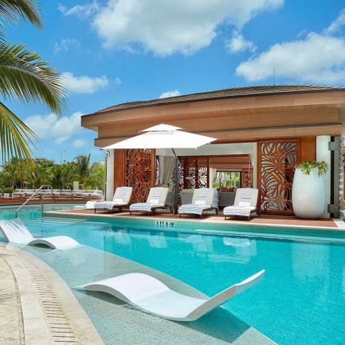 A serene pool area with lounge chairs, an umbrella, and a stylish cabana surrounded by palm trees and blue skies.