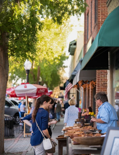 A bustling street market with people browsing wooden crafts and enjoying the vibrant atmosphere under green trees.