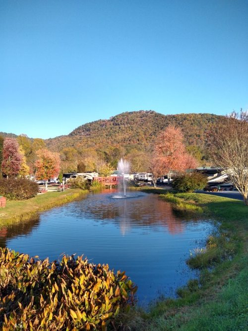 A serene pond with a fountain, surrounded by autumn trees and mountains under a clear blue sky.