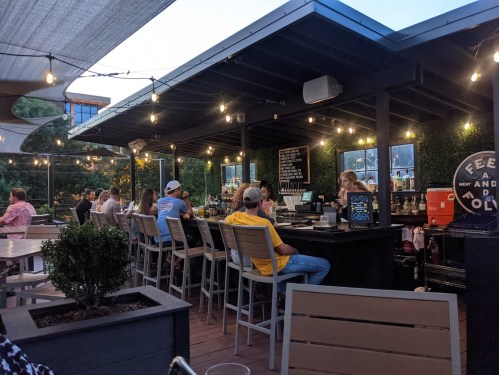 A lively outdoor bar scene with patrons seated at high tables, string lights overhead, and a bartender serving drinks.