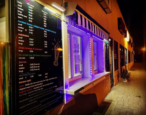 A brightly lit ice cream shop at night, featuring a menu board and colorful lights along the windows.