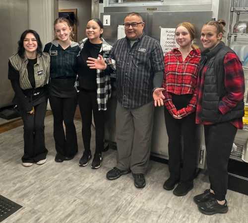 A group of six people, including four women in black outfits and two in plaid shirts, pose together in a kitchen setting.