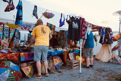 A vibrant market stall with colorful textiles, clothing, and crafts, featuring people browsing and interacting under a tent.