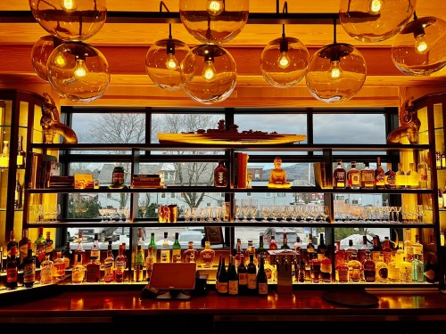 A well-lit bar with shelves of various bottles, glasses, and a view of the outdoors through large windows.