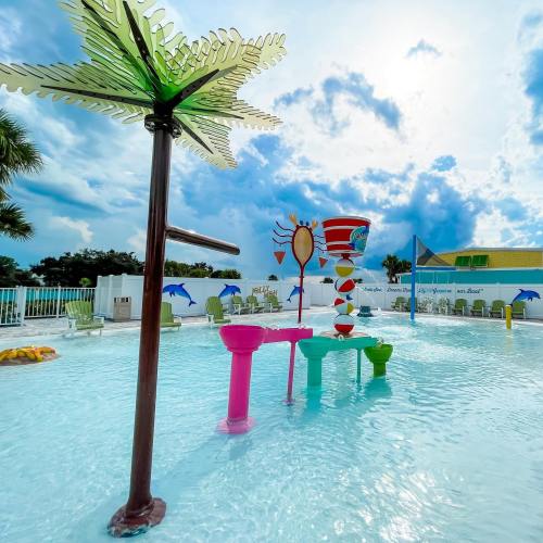 Colorful water play area with palm trees, a large drink fountain, and vibrant seating under a bright sky.
