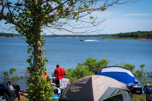 A serene lakeside scene with a man cooking, tents set up, and a boat speeding across the water in the background.