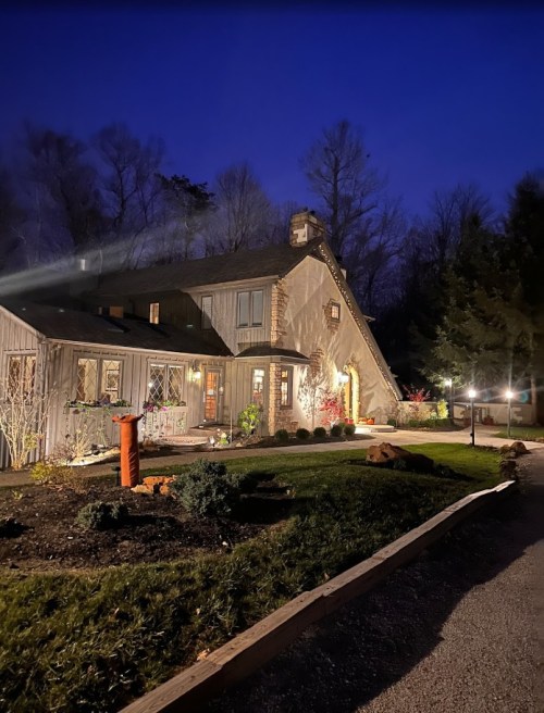 A cozy house at night, illuminated by soft lights, surrounded by trees and landscaped gardens.