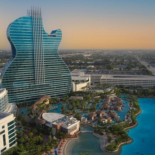 Aerial view of a guitar-shaped hotel with a large pool and lush landscaping at sunset.