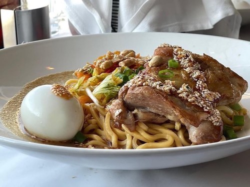 A plate of noodles topped with sesame chicken, a soft-boiled egg, and garnished with vegetables and sesame seeds.