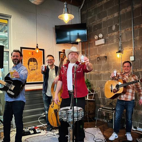 A band of four musicians holds drinks while performing in a cozy venue with a rustic backdrop.
