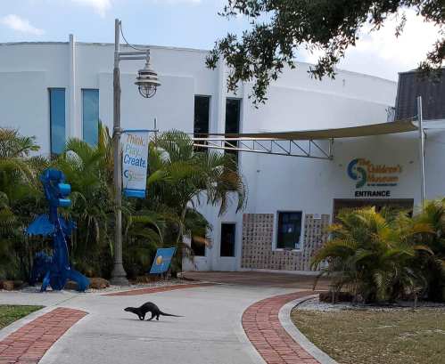 A pathway leads to the entrance of The Children's Museum, with a sculpture and palm trees lining the way. An otter crosses the path.