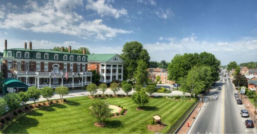 A panoramic view of a historic building with green lawns and trees, alongside a road and blue sky.
