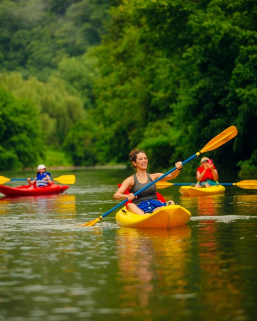 A woman paddles a yellow kayak on a calm river, surrounded by lush greenery and other kayakers in the background.