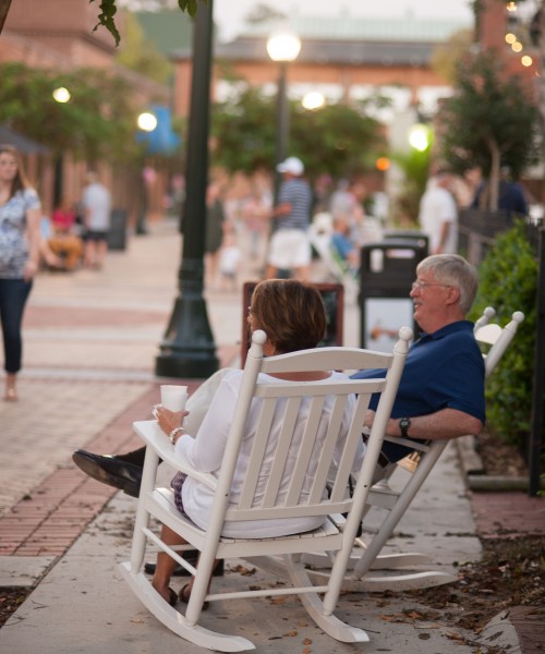 A couple relaxes in rocking chairs on a bustling street, enjoying the evening atmosphere.