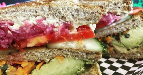 A close-up of a colorful sandwich with layers of vegetables, served on whole grain bread, against a vibrant chalkboard menu.
