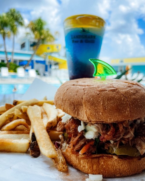 A pulled pork sandwich with pickles and coleslaw, served with fries and a colorful drink, by a poolside setting.