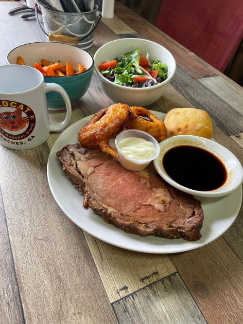 A plate of prime rib with sides of onion rings, salad, and carrots, alongside a mug of coffee on a wooden table.