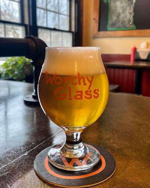 A glass of beer with a frothy top sits on a coaster, featuring the text "Worthy Glass" in bright orange.