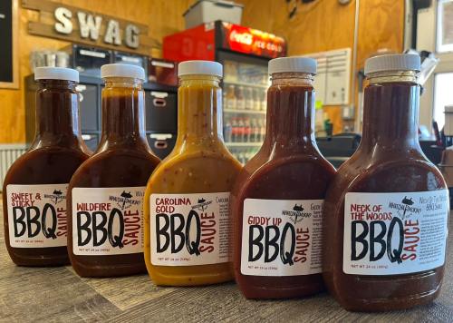 Five bottles of BBQ sauce lined up on a counter, each with distinct labels and flavors, in a rustic setting.