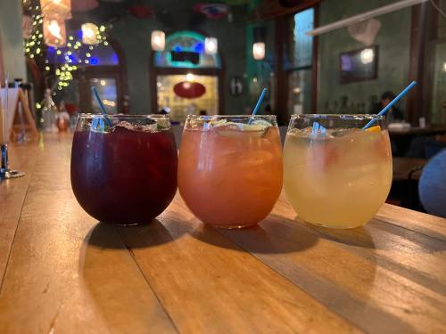 Three colorful cocktails in round glasses sit on a wooden table, with a cozy bar atmosphere in the background.