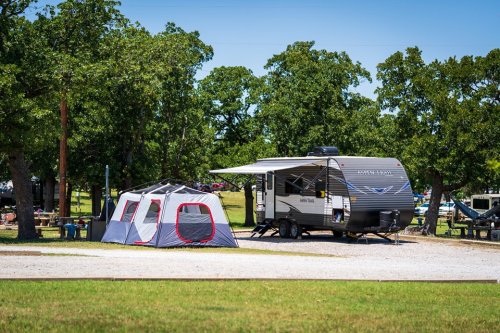 A campsite featuring a travel trailer and a tent, surrounded by green trees and open grass.