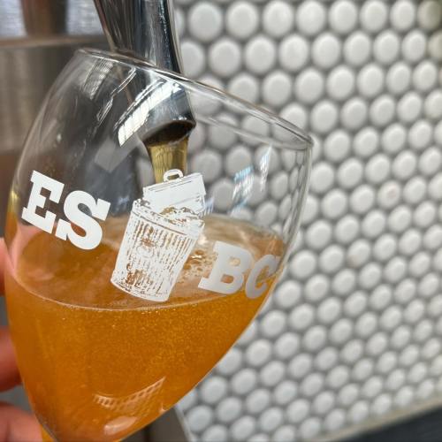 A close-up of a glass being filled with a golden beverage, featuring a logo with a trash can design.