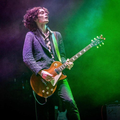 A musician with curly hair plays an electric guitar on stage, surrounded by colorful lighting.