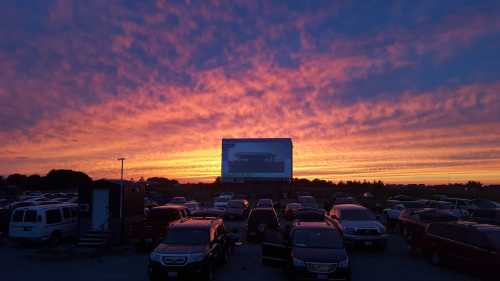 A drive-in movie theater at sunset, with colorful clouds and parked cars in the foreground.