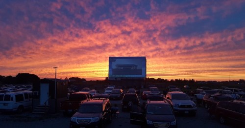 A vibrant sunset with colorful clouds behind a drive-in movie screen and parked cars in the foreground.