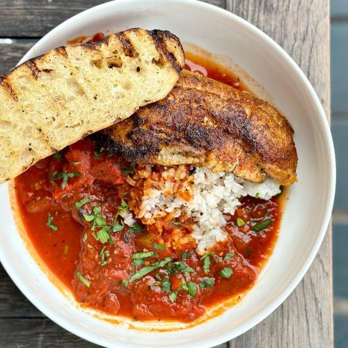 A bowl of rice topped with a spicy tomato sauce and a piece of grilled fish, served with a slice of toasted bread.