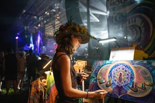 An artist paints on a canvas at a vibrant outdoor festival, surrounded by colorful lights and a lively crowd.